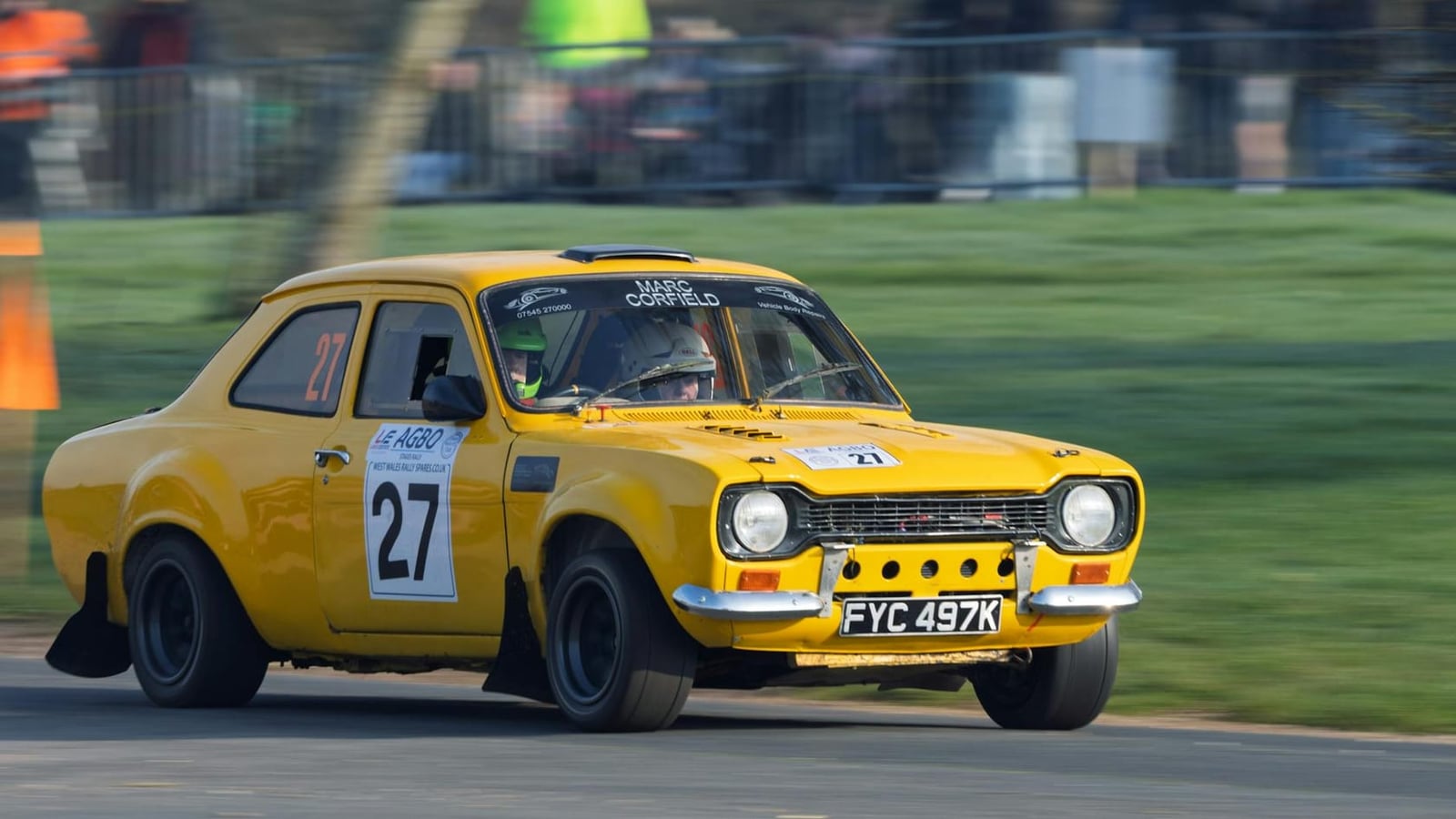 A yellow Ford Escort Mk1 rally car with competition number 27 cornering hard on a stage