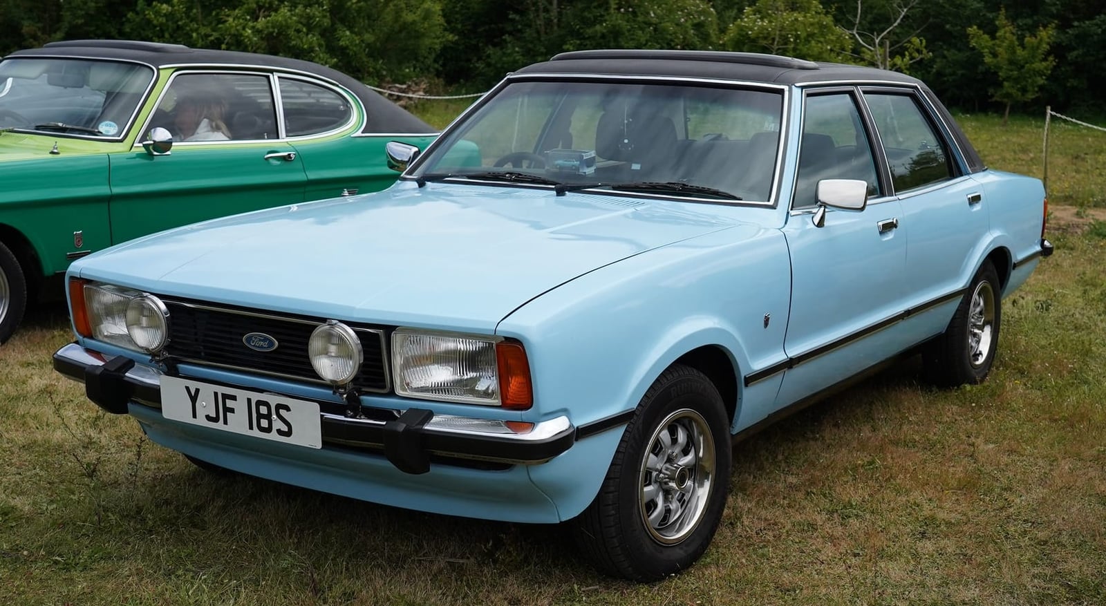 A light blue Ford Cortina Ghia four-door saloon with a vinyl roof, front three-quarter view at a show